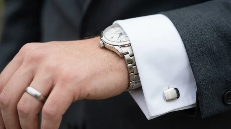 תקריב של יד גבר עונדת טבעת נישואין כסופה, שעון יד, וחפתים כסופים על חפת חולצה לבנה וחליפה אפורה כהה.